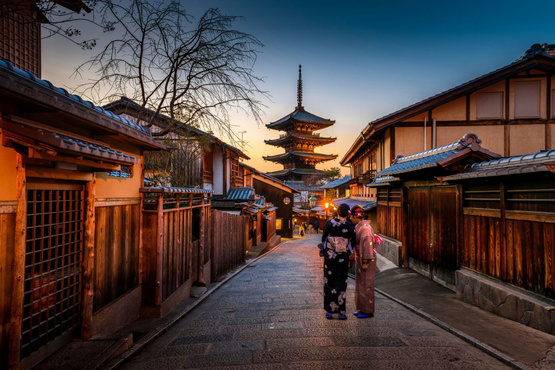 Two women in kimonos walking a traditional Kyoto street at dusk with Yasaka Pagoda in the distance